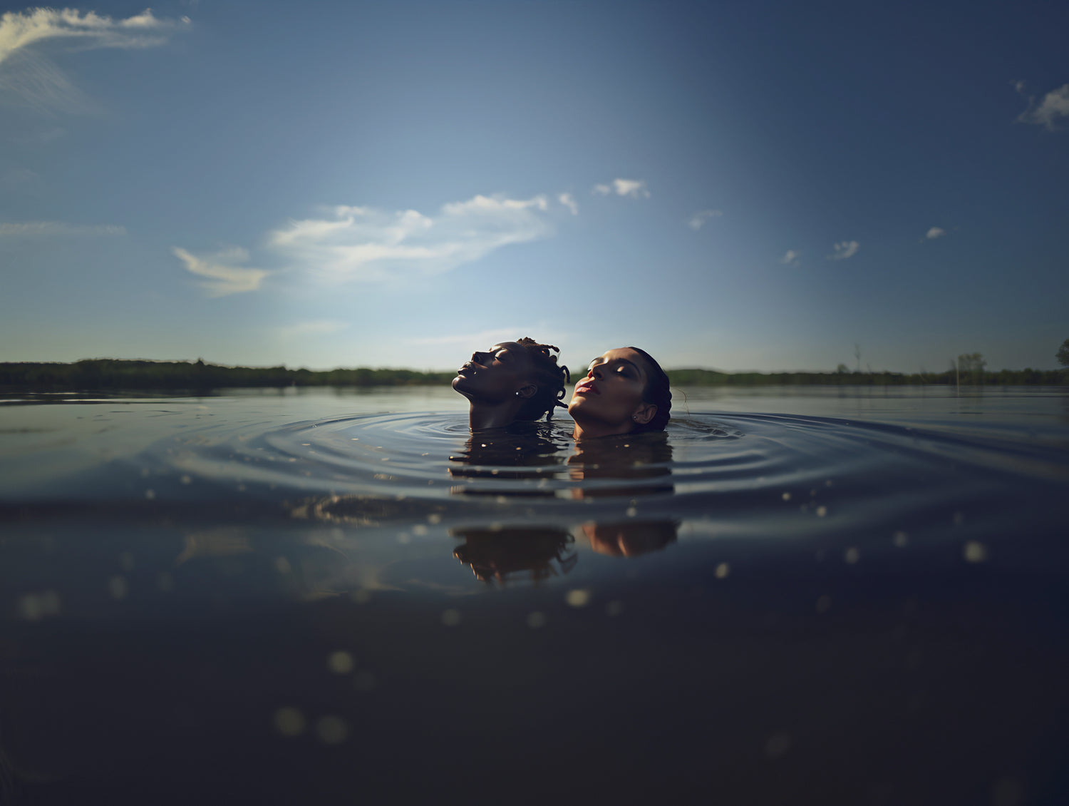 A wide angle image of two beautiful models submerged in a lake with only their faces sticking out of the water with a beautiful sky behind them.